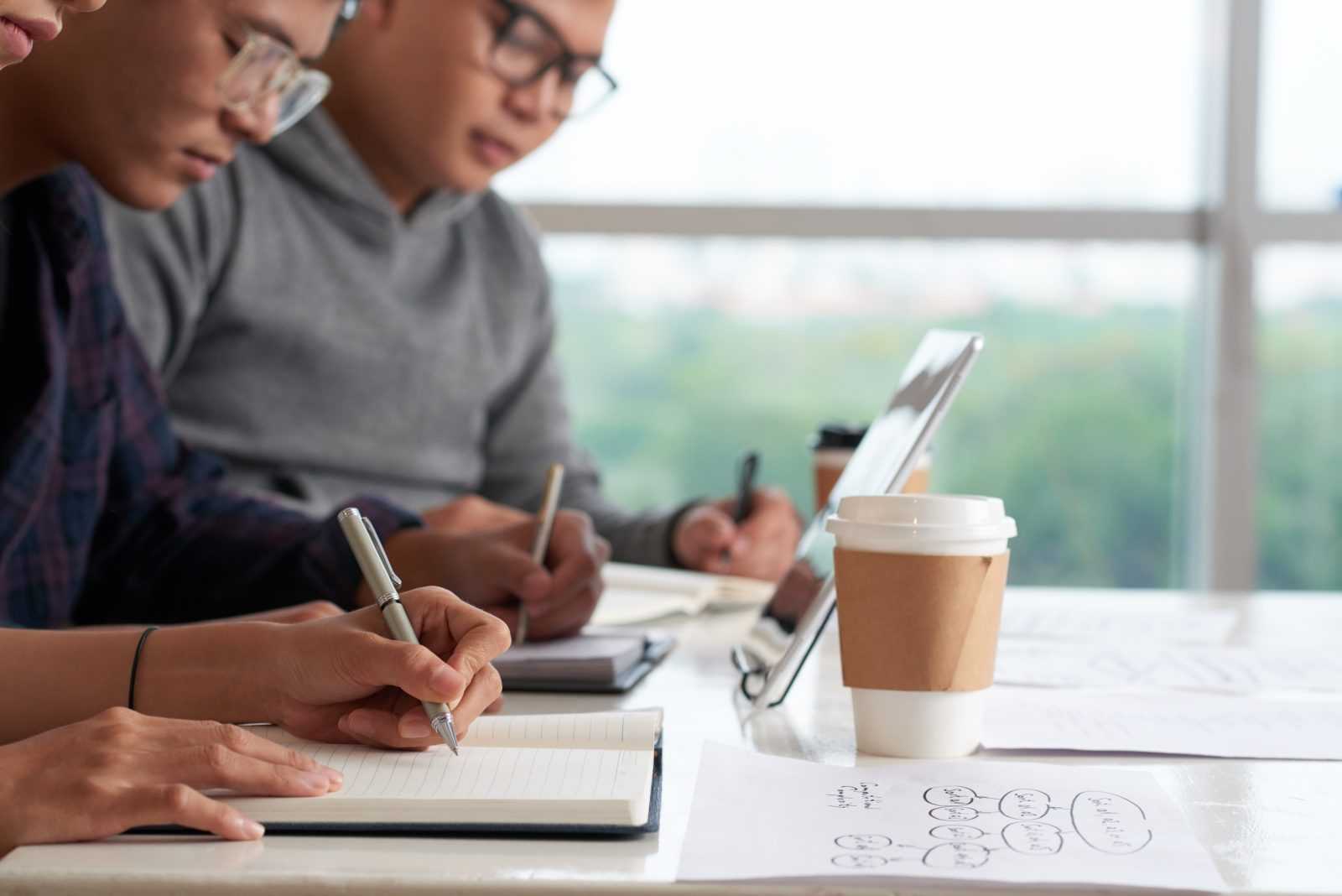 Close-up shot of concentrated Asian students sitting at wooden desk and taking necessary notes while having lecture